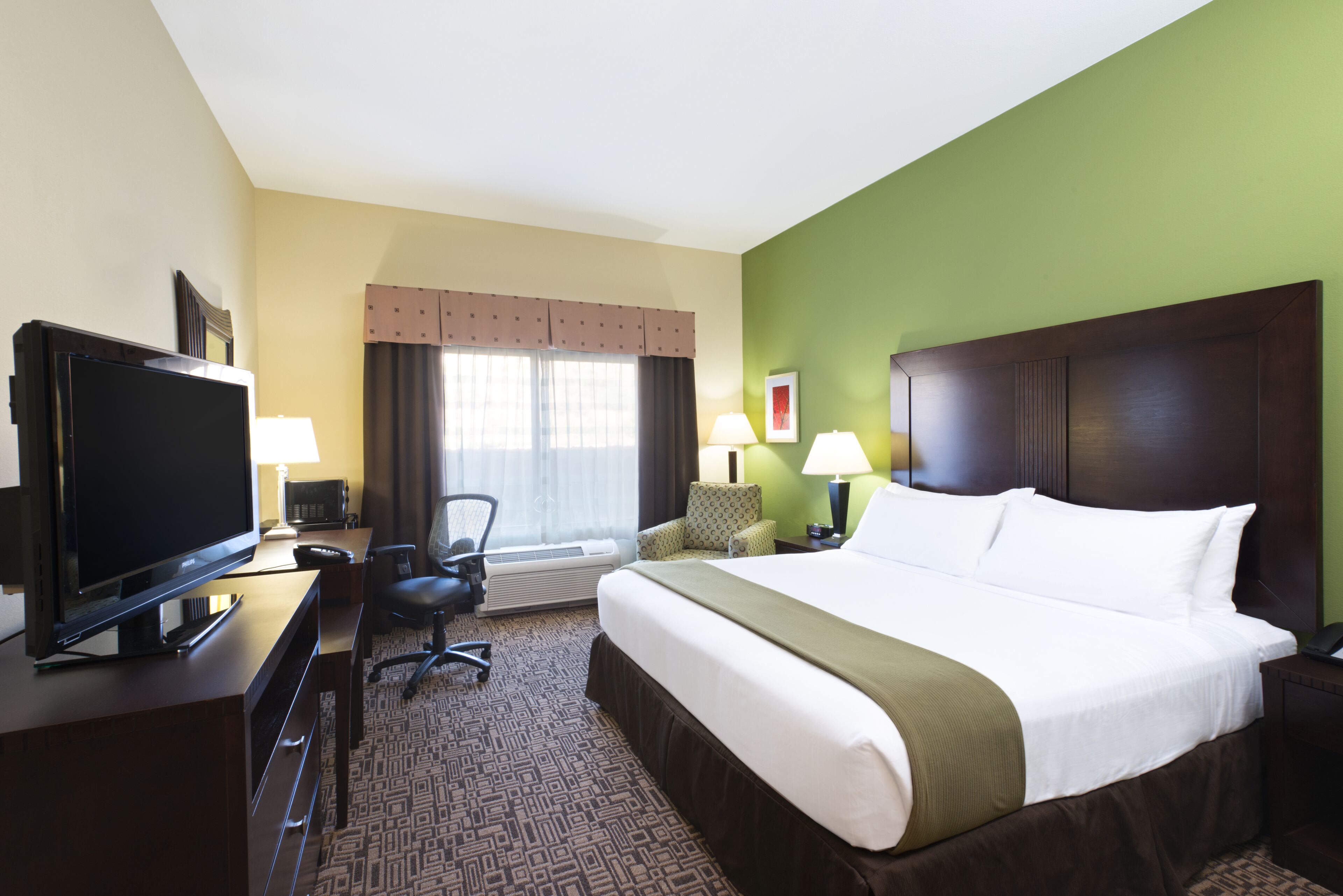 A hotel room with a large bed, a television, a desk with a computer, and two lamps.