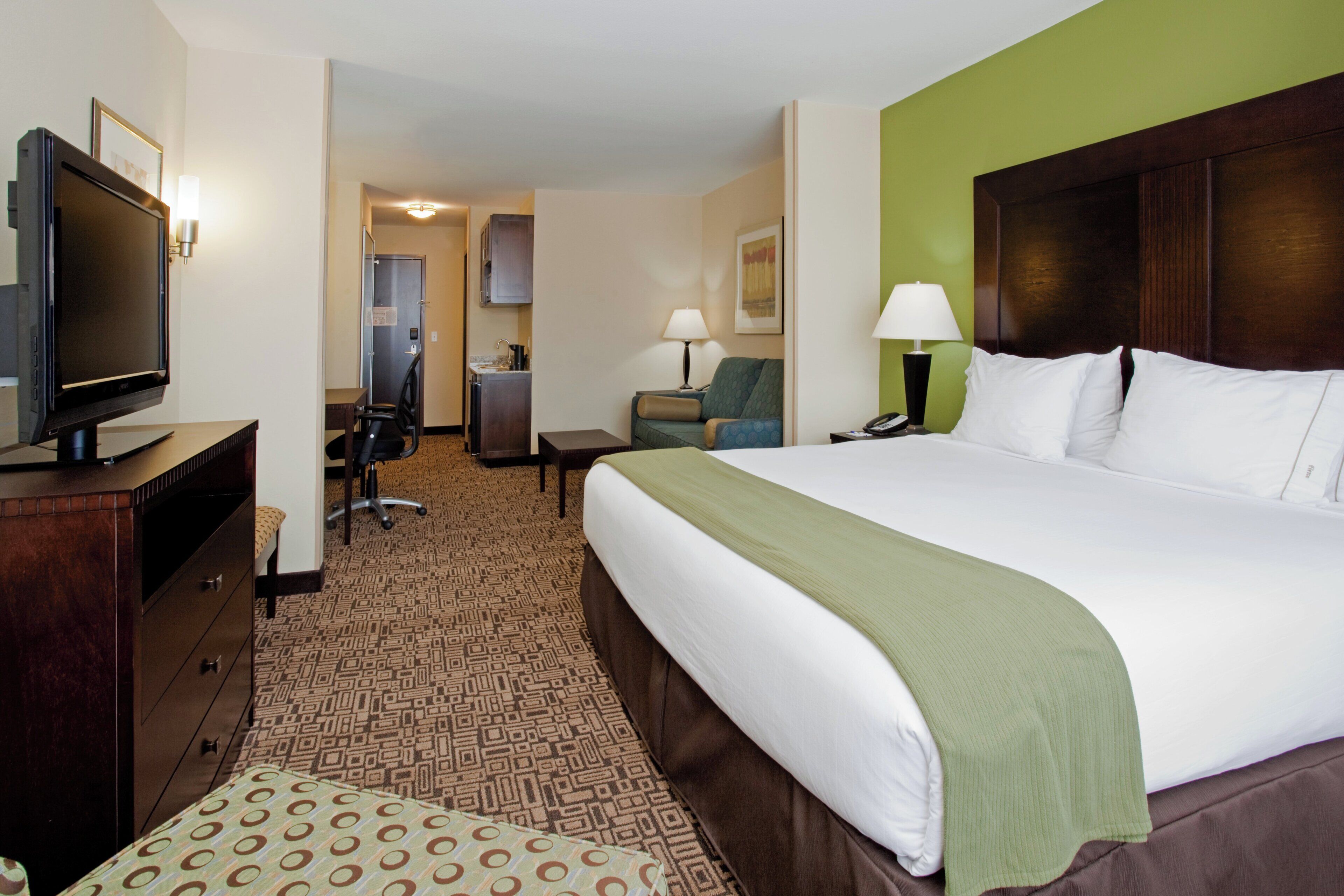 A hotel room with a large bed, a desk with a television, a chair, a lamp, and a carpeted floor.