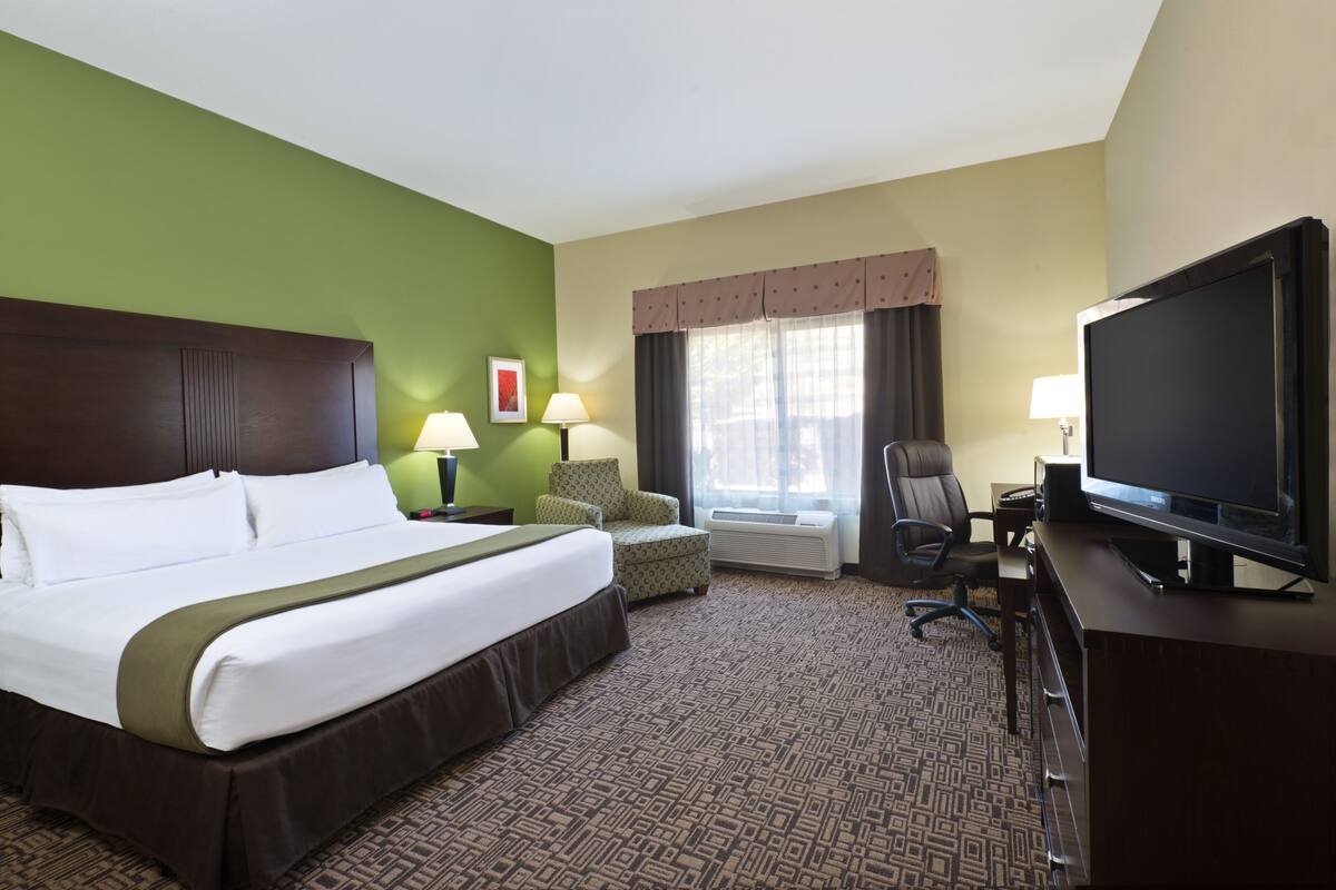 A hotel room with a large bed, a desk with a television, a chair, a desk lamp, and a window with curtains.