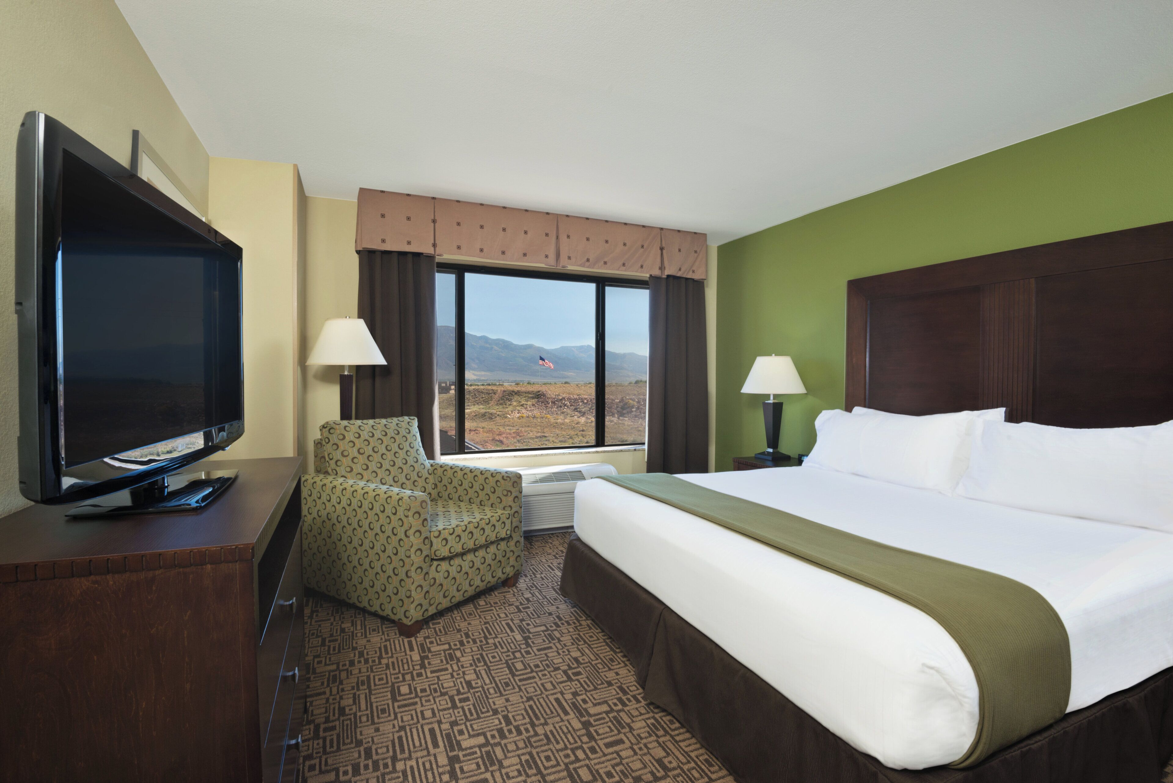 A hotel room with a large bed, a flat-screen TV, a patterned armchair, a window with a view of mountains, and a green wall.