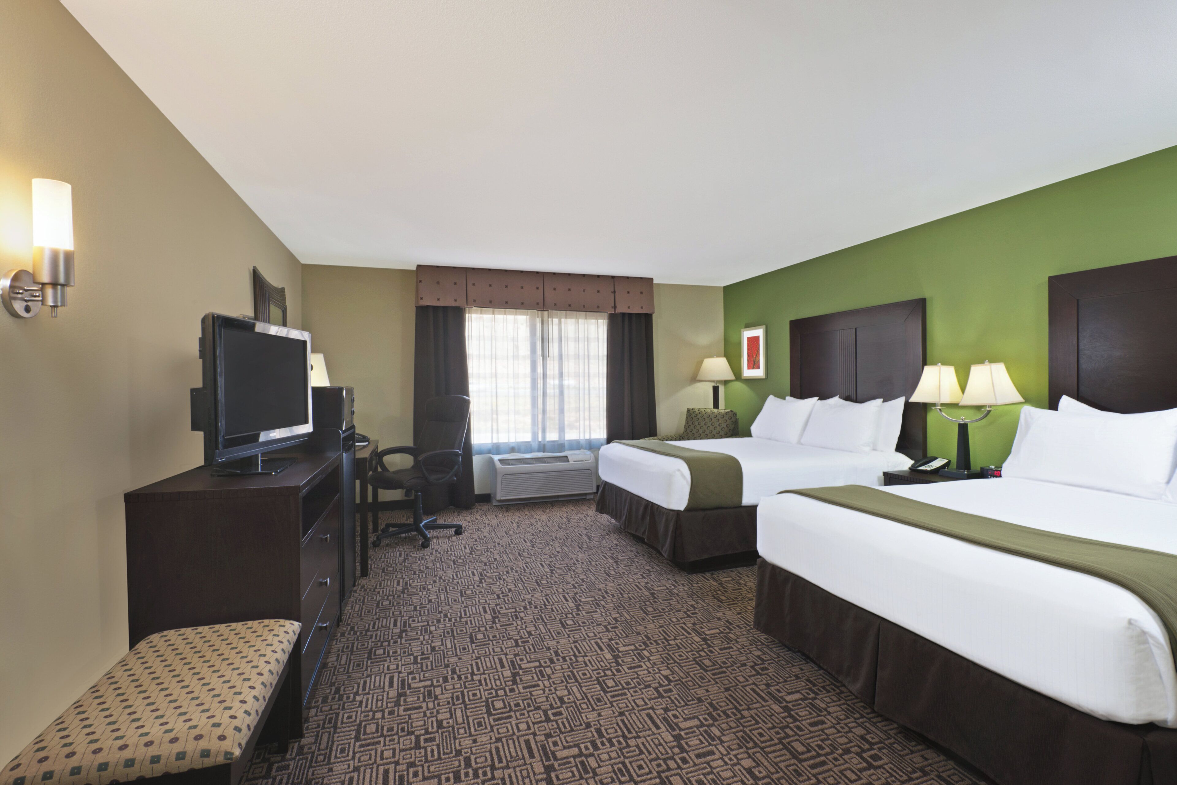 A hotel room with a bed, a desk with a television, a chair, and a window with curtains.