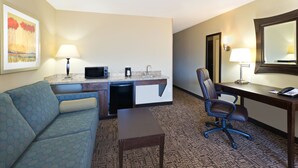 A hotel room with a sofa, a desk, a chair, a coffee maker, a microwave, and a painting on the wall.