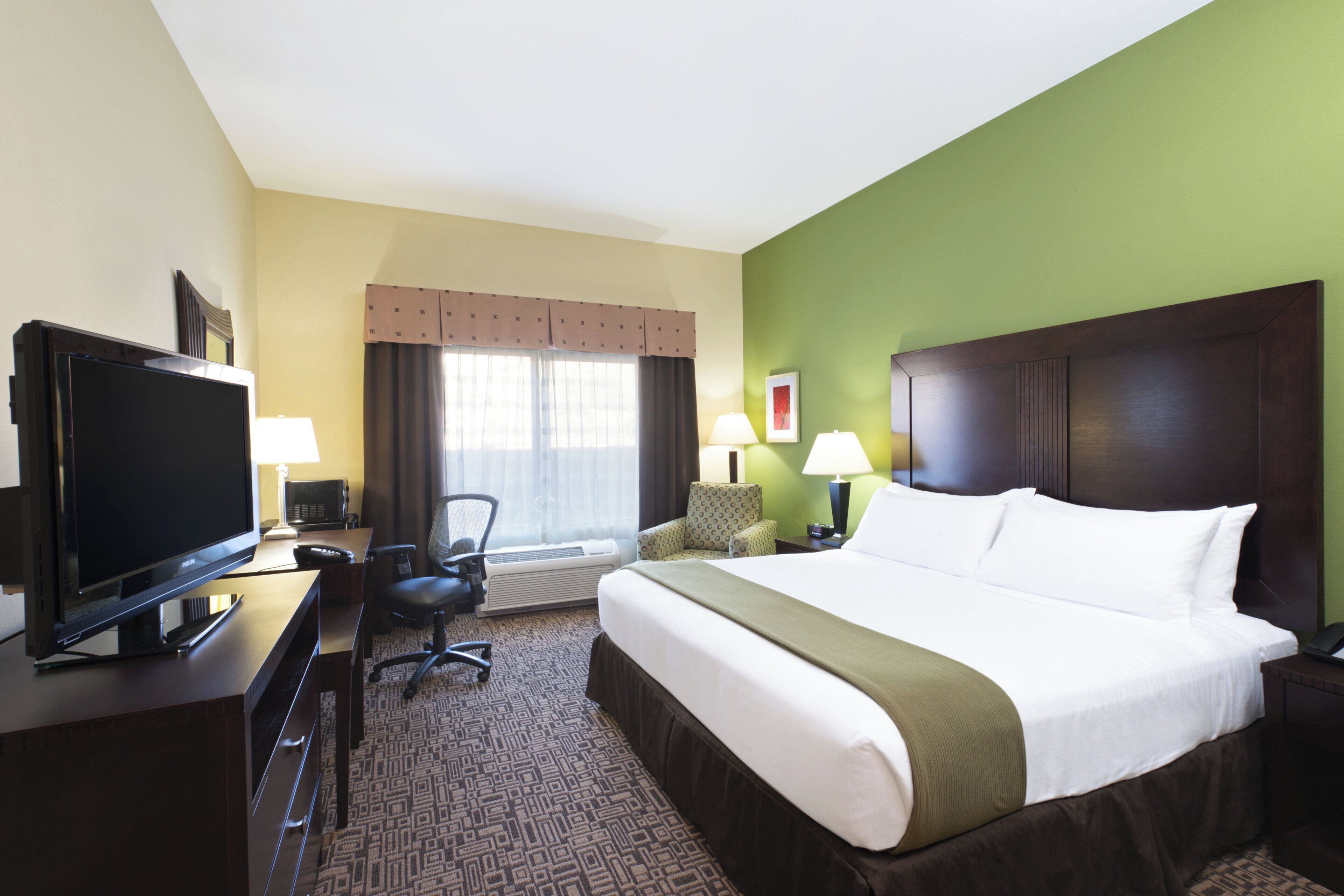 A hotel room with a bed, television, desk, chair, lamps, and a window with curtains.