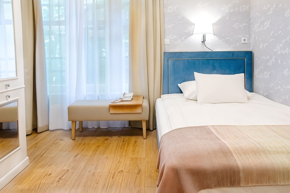 Una habitación de hotel con cama, mesita de noche, lámpara y ventana con cortinas.