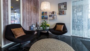 Una zona de recepción moderna con dos sillas negras, una mesa de vidrio, una lámpara decorativa y una pared con cuadros y una planta.