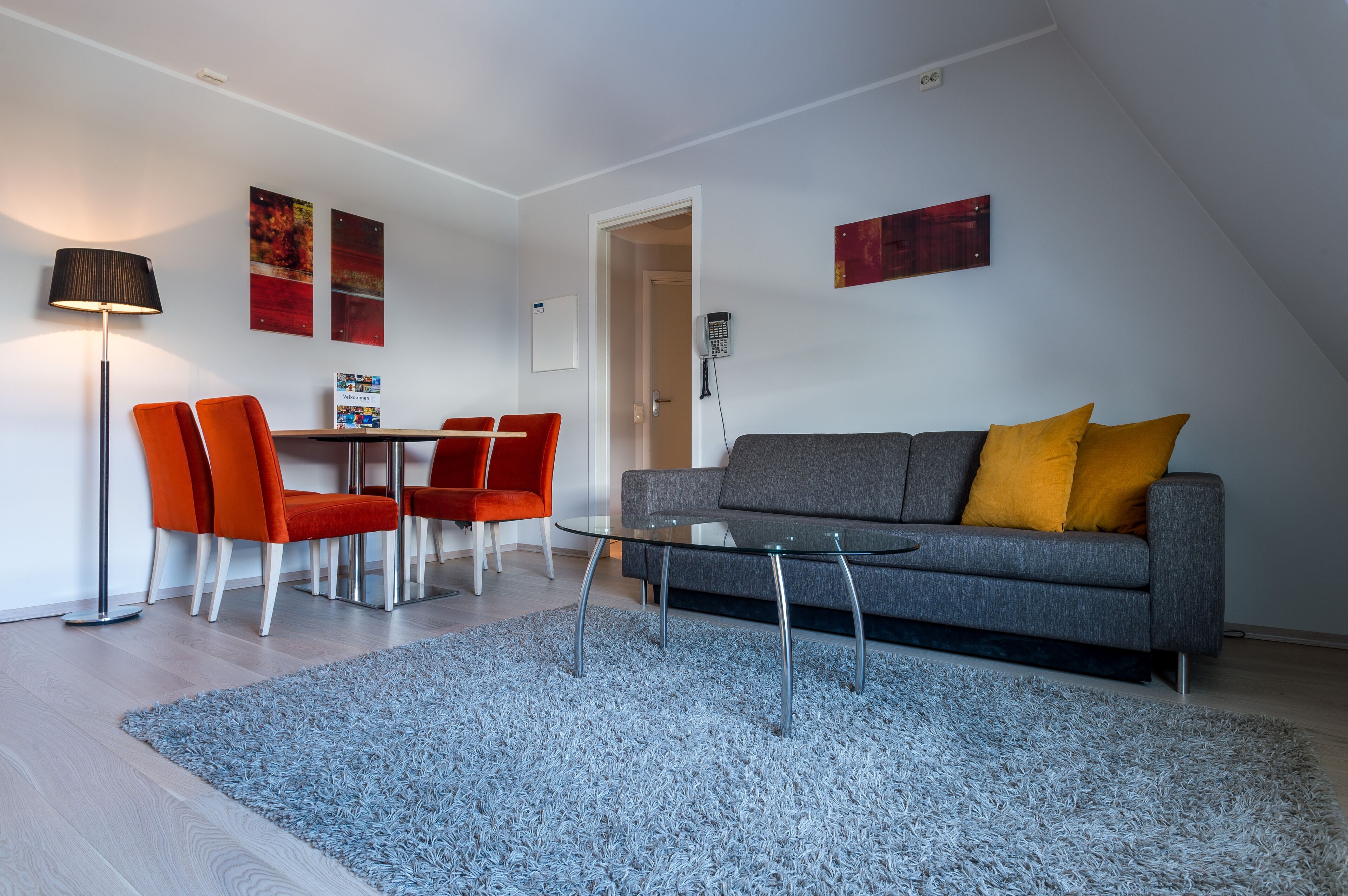 A modern living room with a grey sofa, orange chairs, a glass coffee table, and a blue shag rug.