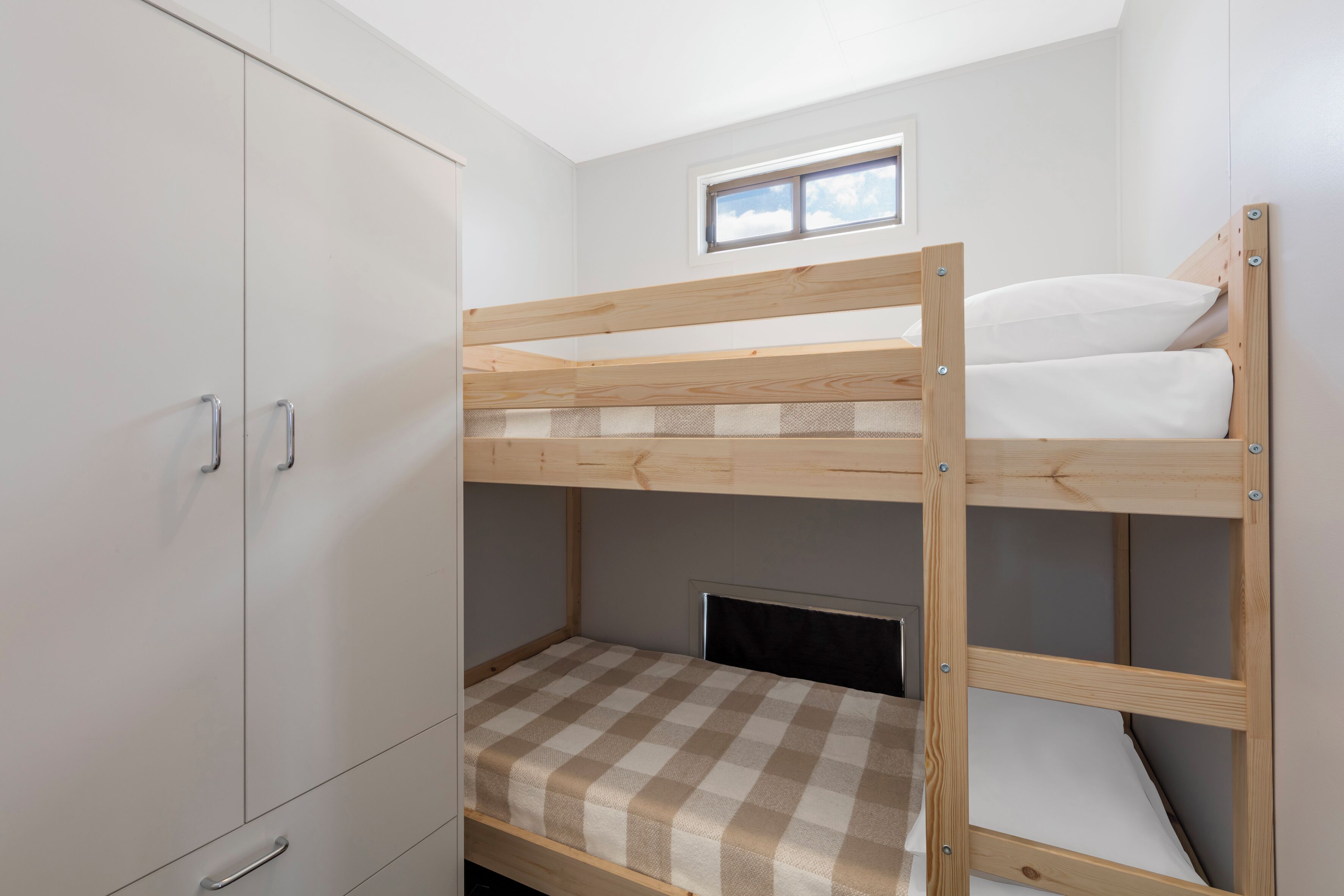 Een stapelbed met een geruit matras, een klein raam erboven en een kledingkast met twee deuren.