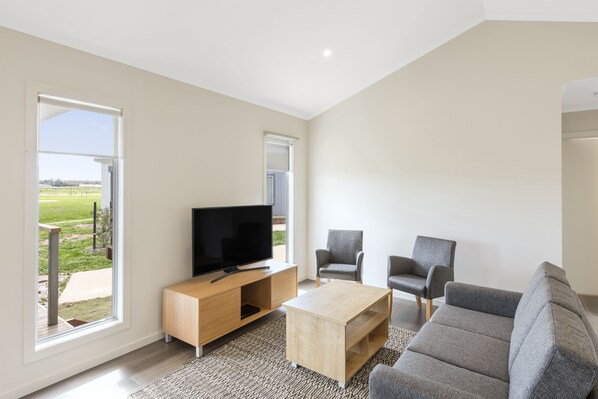 A modern living room with a flat-screen TV, two grey armchairs, a wooden coffee table, and large windows offering a view of a green landscape.