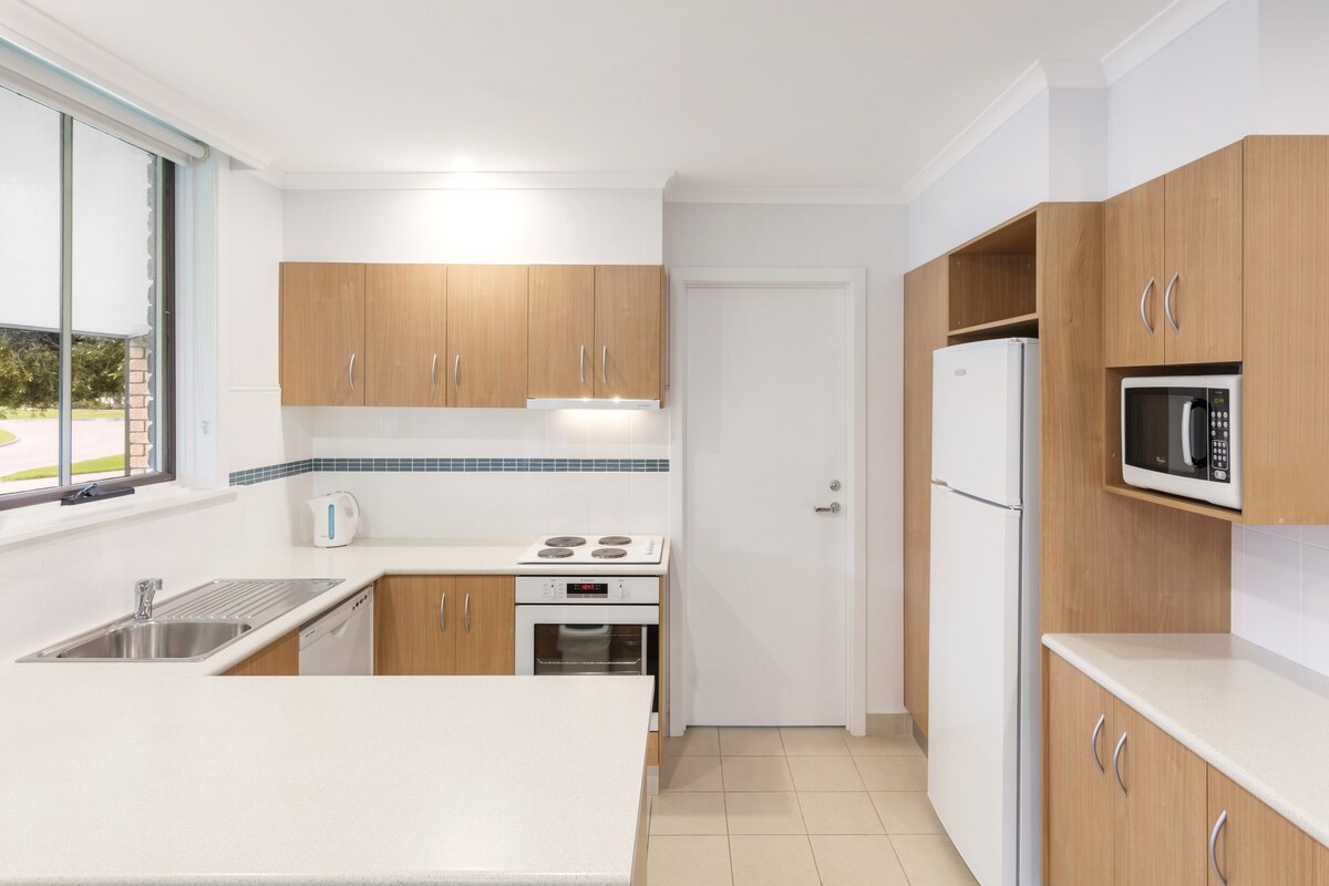 A modern kitchen with wooden cabinets, a white countertop, a built-in microwave, and a window with a view.