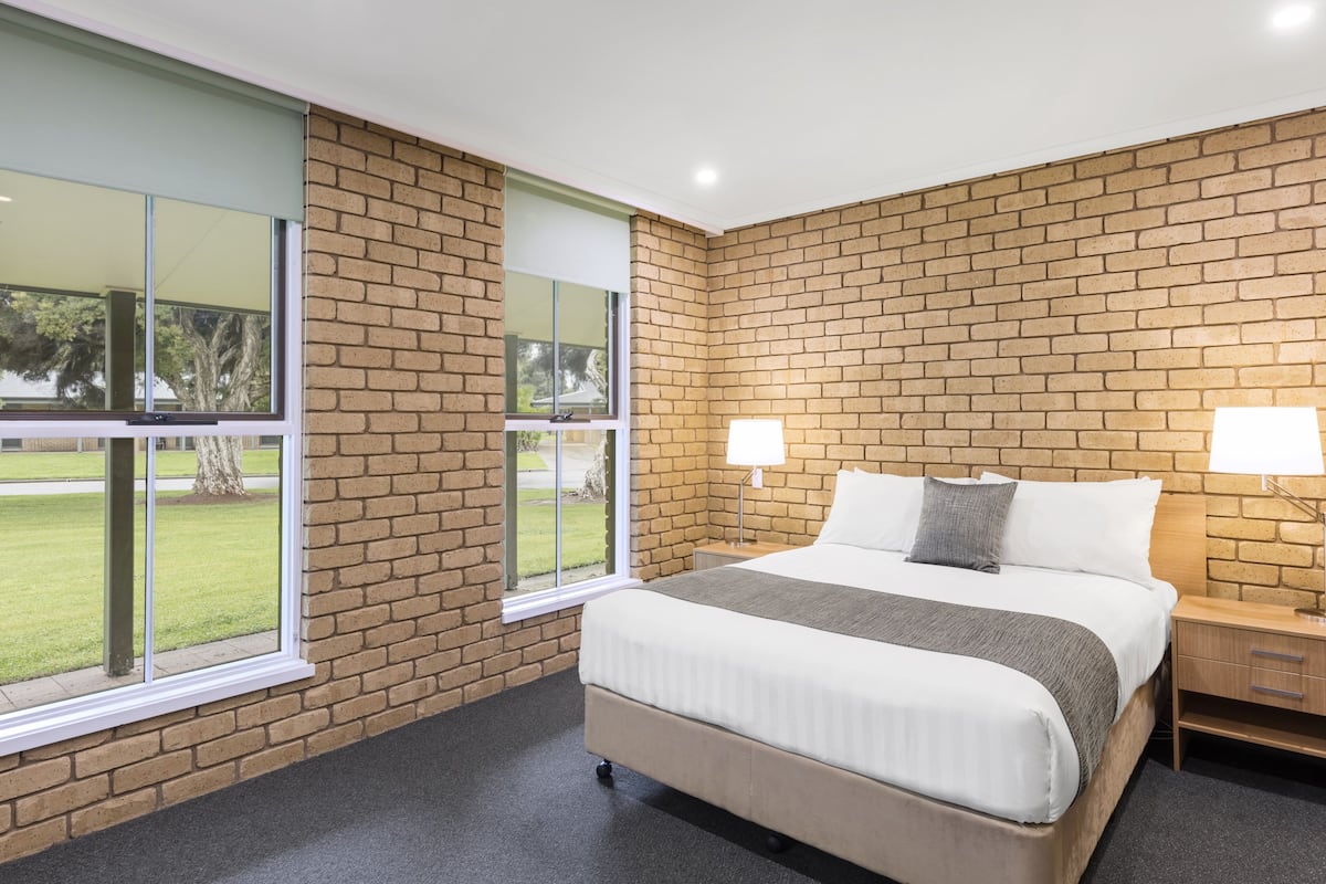 A hotel room with a brick wall, a large bed with white bedding, two bedside lamps, and a view of a grassy area through a window.