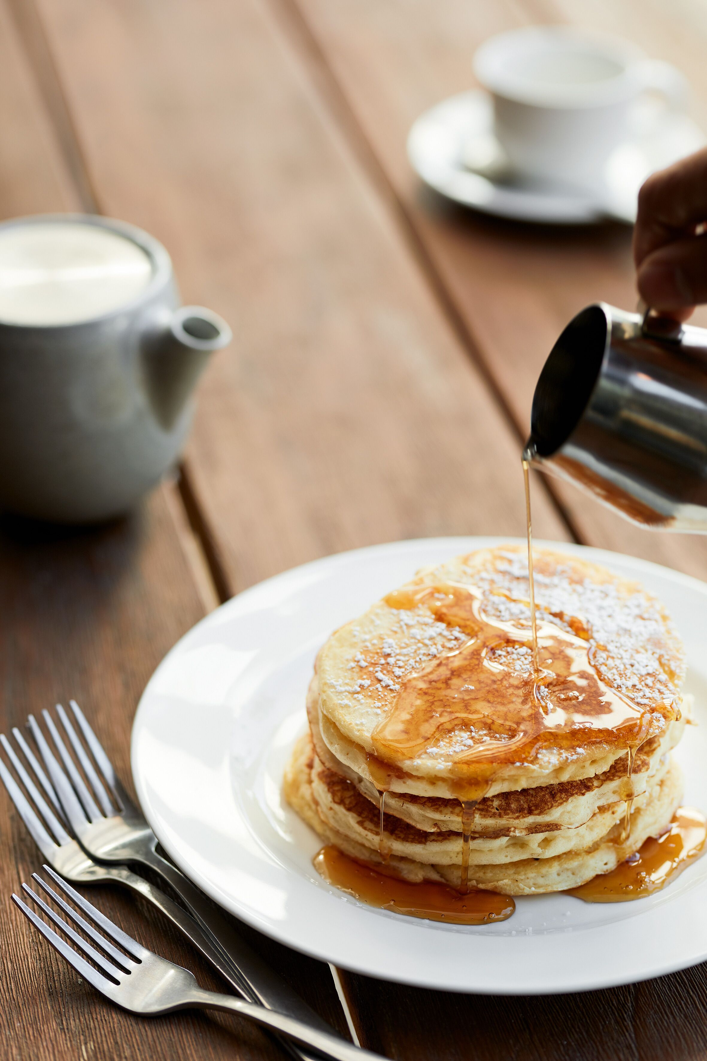 A stack of pancakes on a white plate with syrup being poured over them.