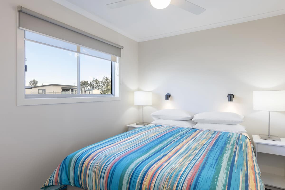 A bedroom with a large window, a bed with a striped bedspread, two bedside lamps, and a ceiling fan.