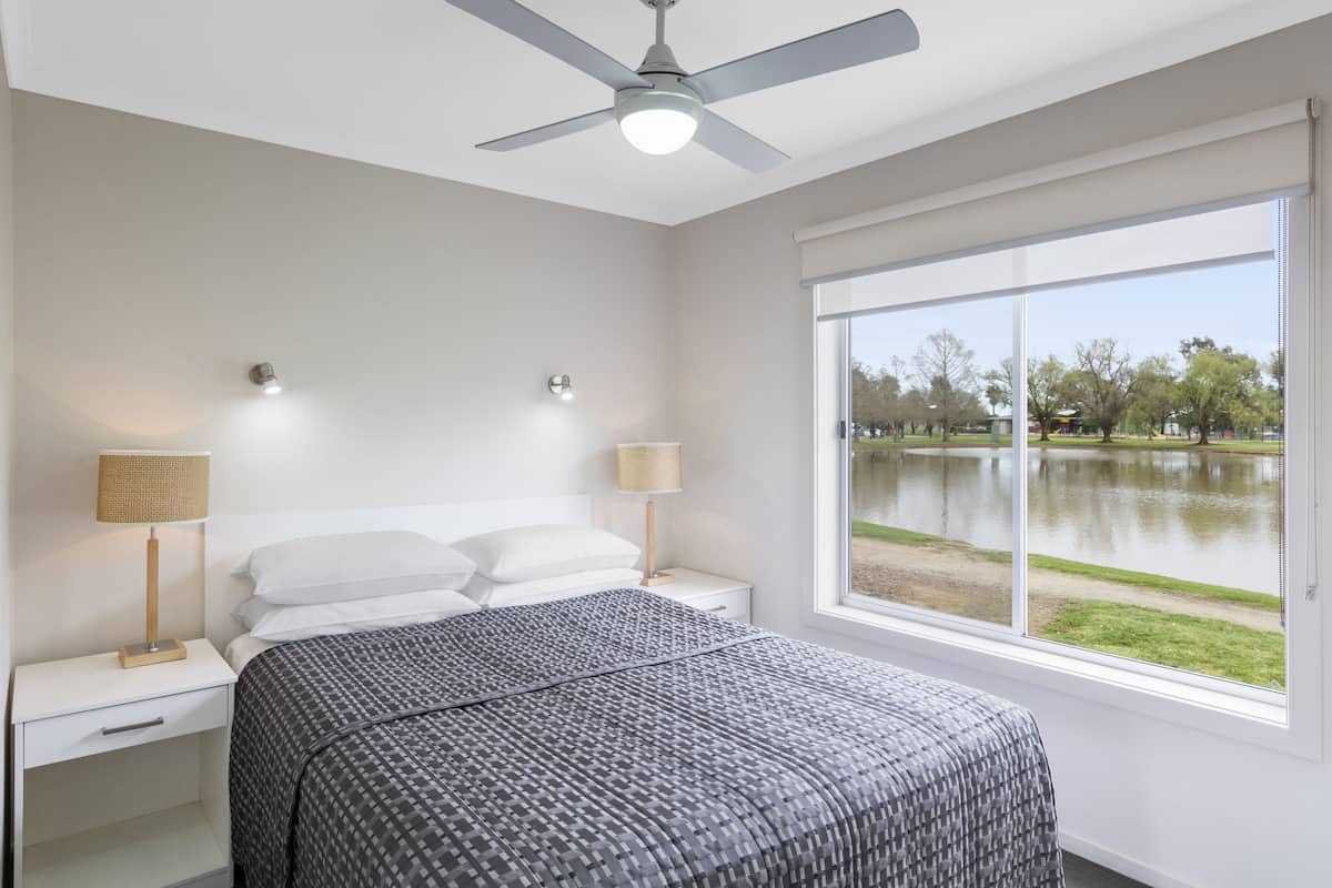 A bedroom with a bed, nightstands, lamps, a ceiling fan, and a large window with a view of a lake and park.