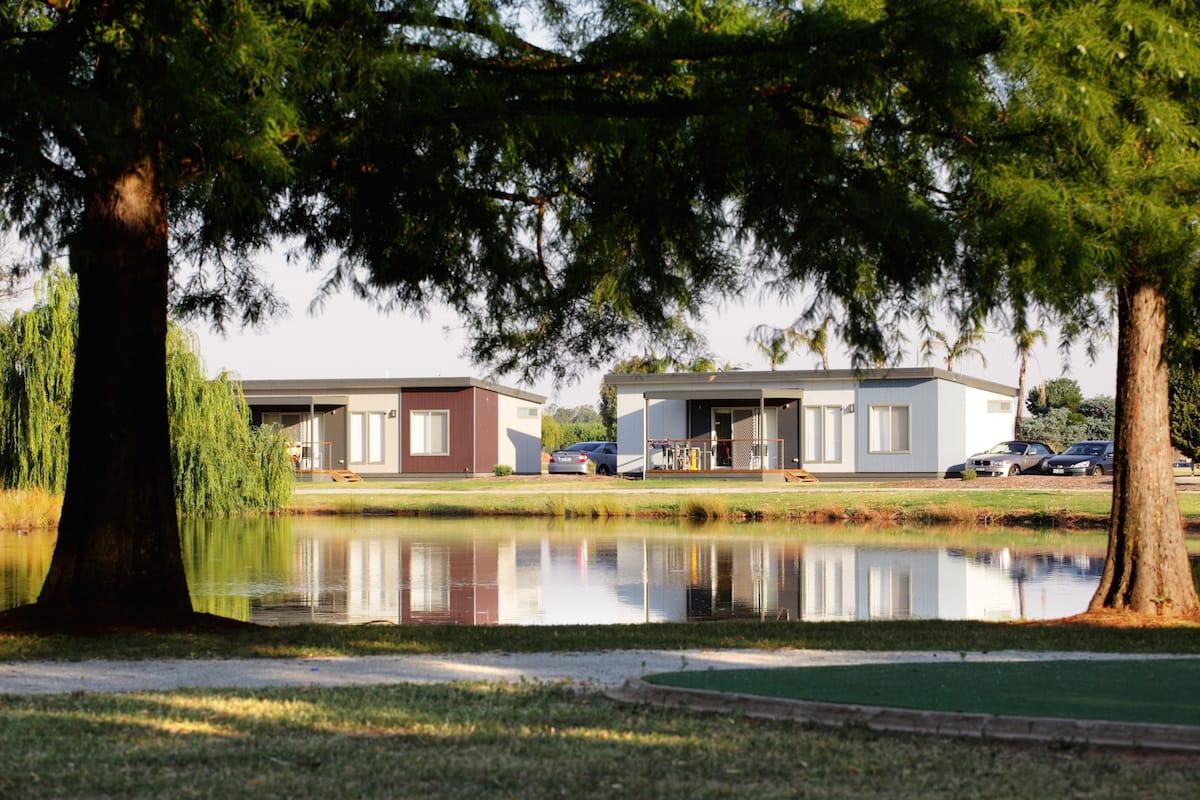A tranquil lakeside setting with two modern buildings, a clear reflection in the water, and a well-maintained grassy area.