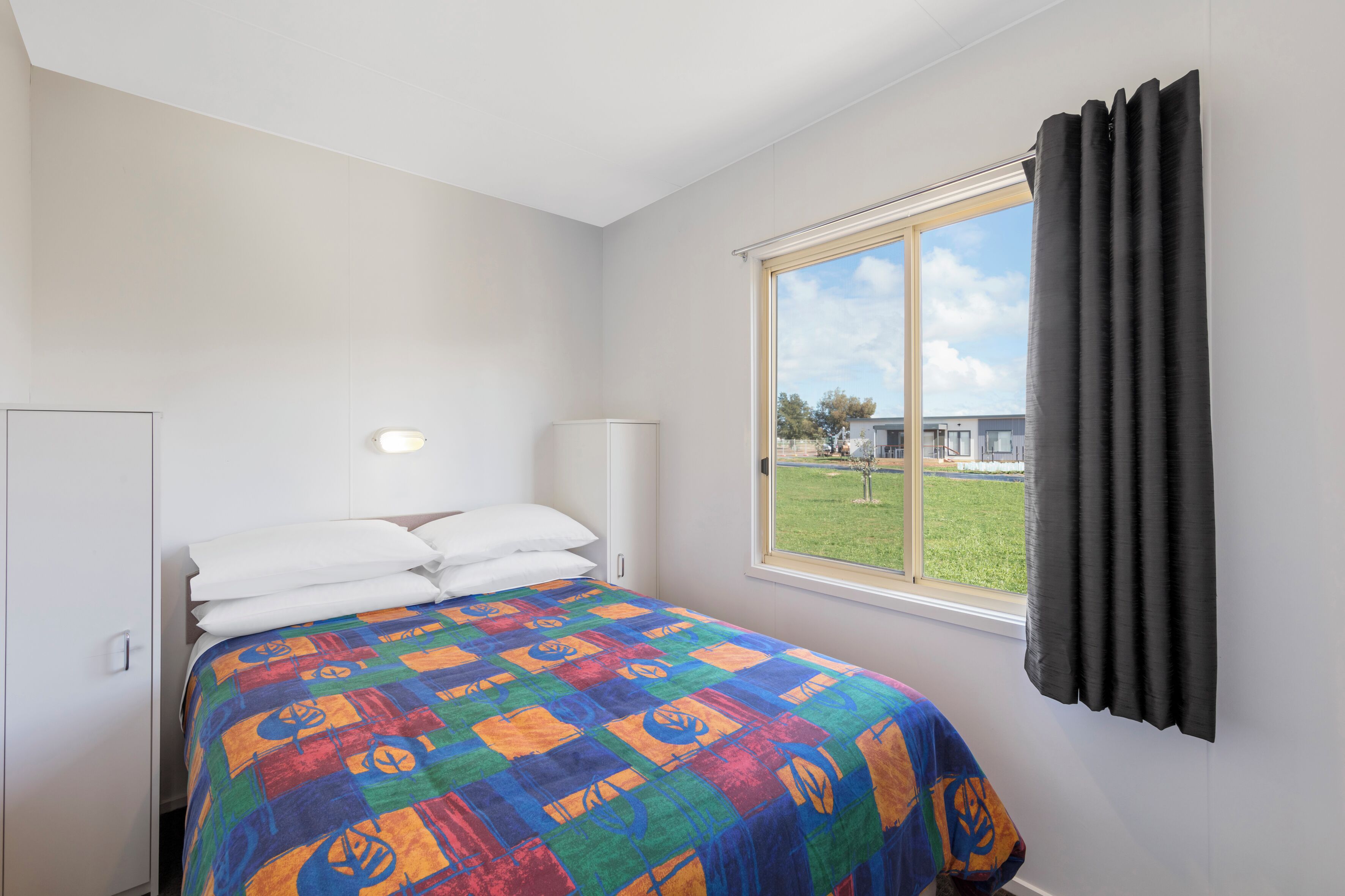 Un dormitorio con cama, ventana con cortinas, lámpara de noche y vista a un espacio verde exterior.
