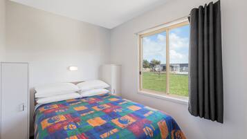 Un dormitorio con cama, ventana con cortinas, lámpara de noche y vista a un espacio verde exterior.