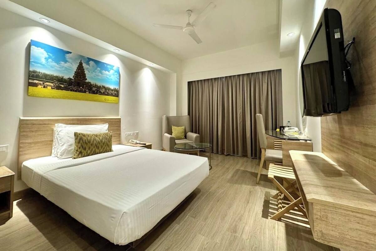 A modern hotel room with a large bed, a desk, a chair, a television, and a painting of a temple.