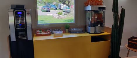 A kitchenette with a coffee machine, a water dispenser, a window with a view of parked cars and greenery, and a cactus plant.