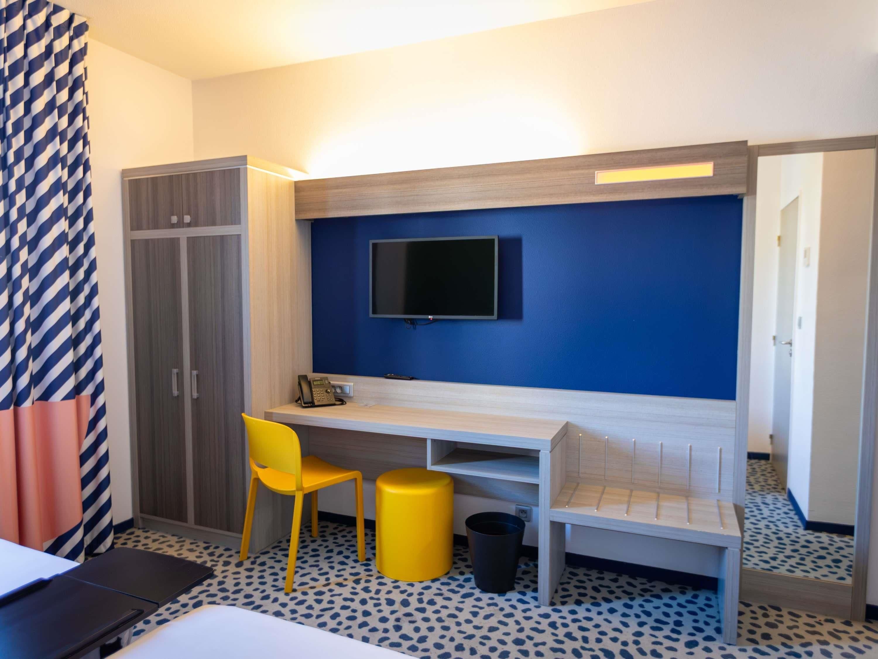 A hotel room with a desk, a chair, a television mounted on the wall, a wardrobe, and a patterned floor.