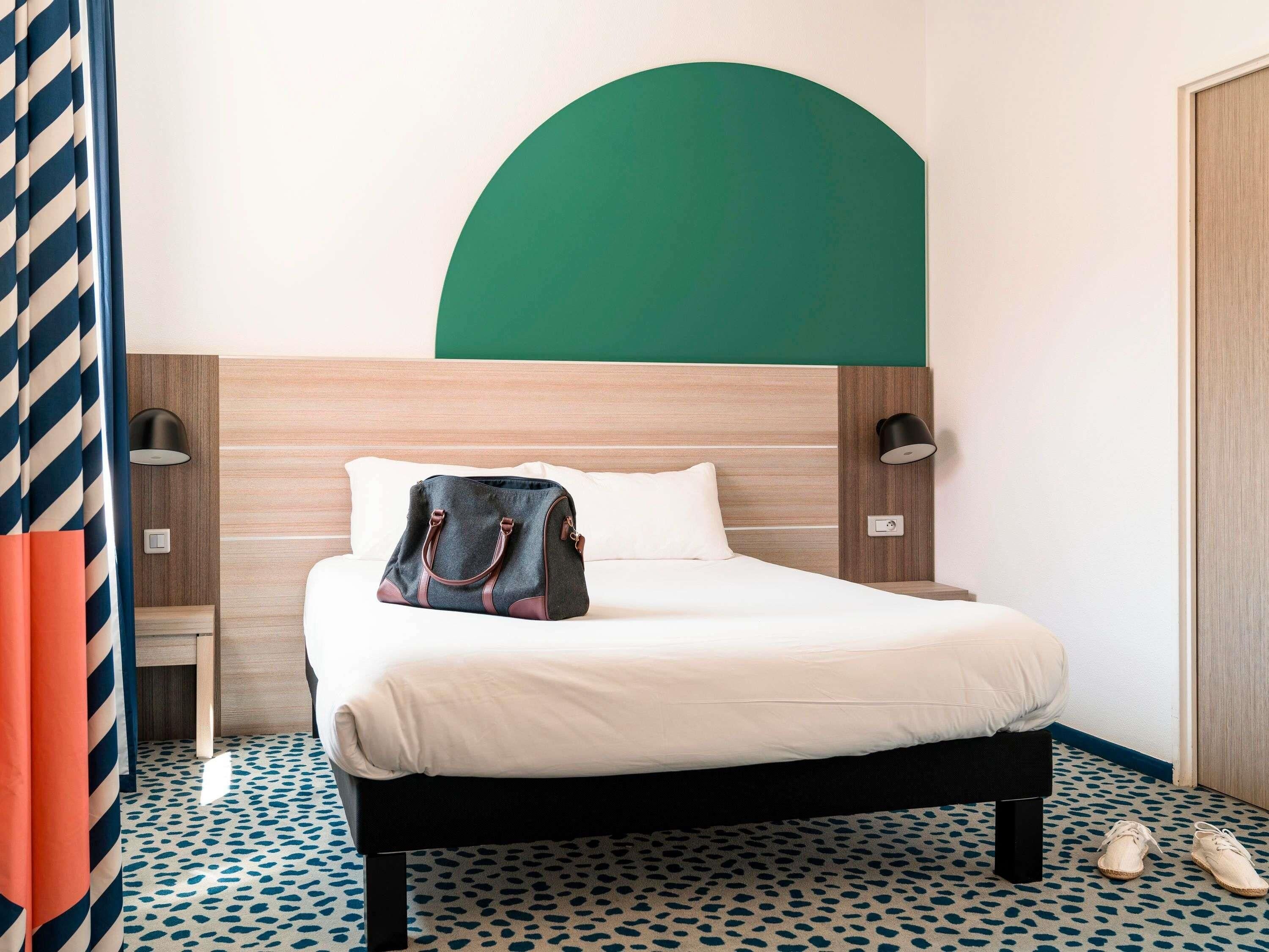 A hotel room with a bed, a nightstand, a lamp, a handbag, and a pair of shoes.
