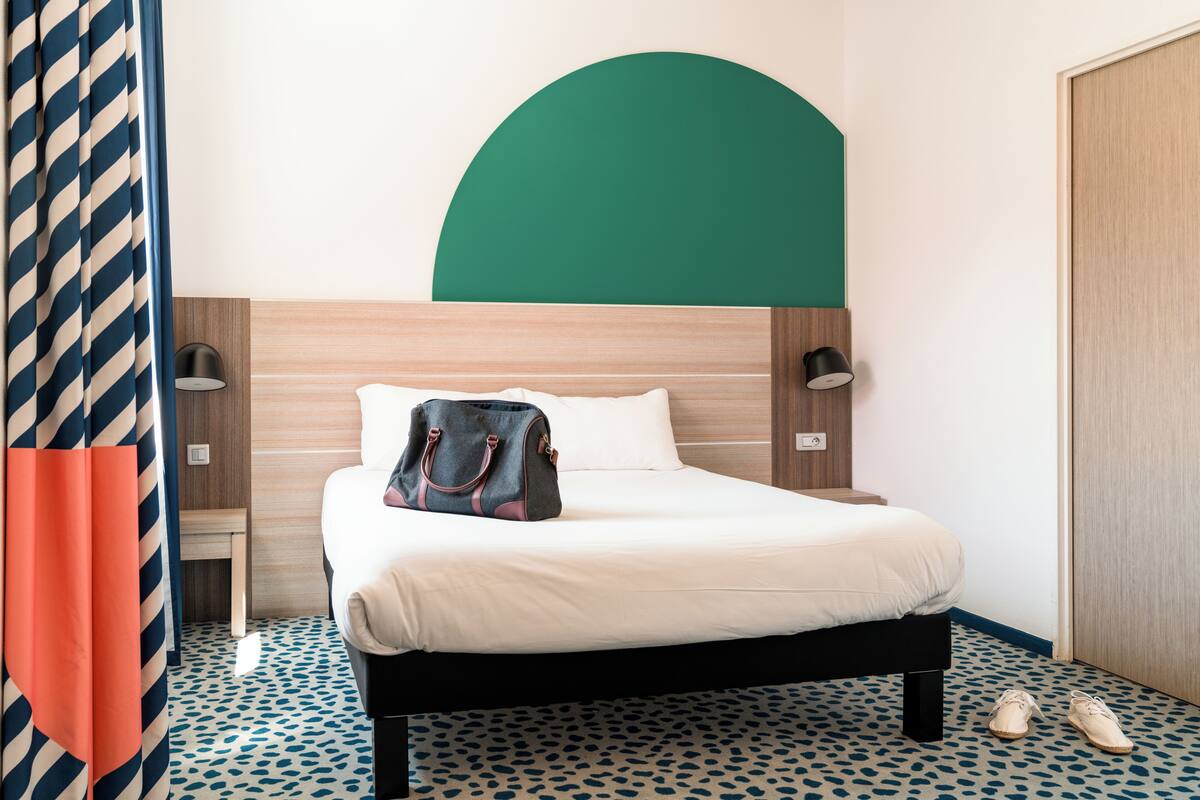 A hotel room with a bed, a nightstand, a lamp, a handbag, and a pair of shoes.