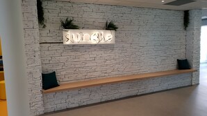 A reception area with a white brick wall, a neon sign reading "Jungle," two dark cushions on a wooden bench, and green plants on the wall.