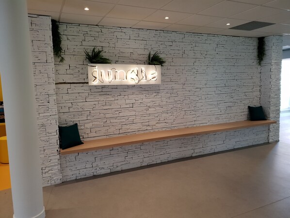 Un área de recepción con pared de ladrillo blanco, un letrero de neón que dice "Jungle", dos cojines oscuros en un banco de madera y plantas verdes en la pared.