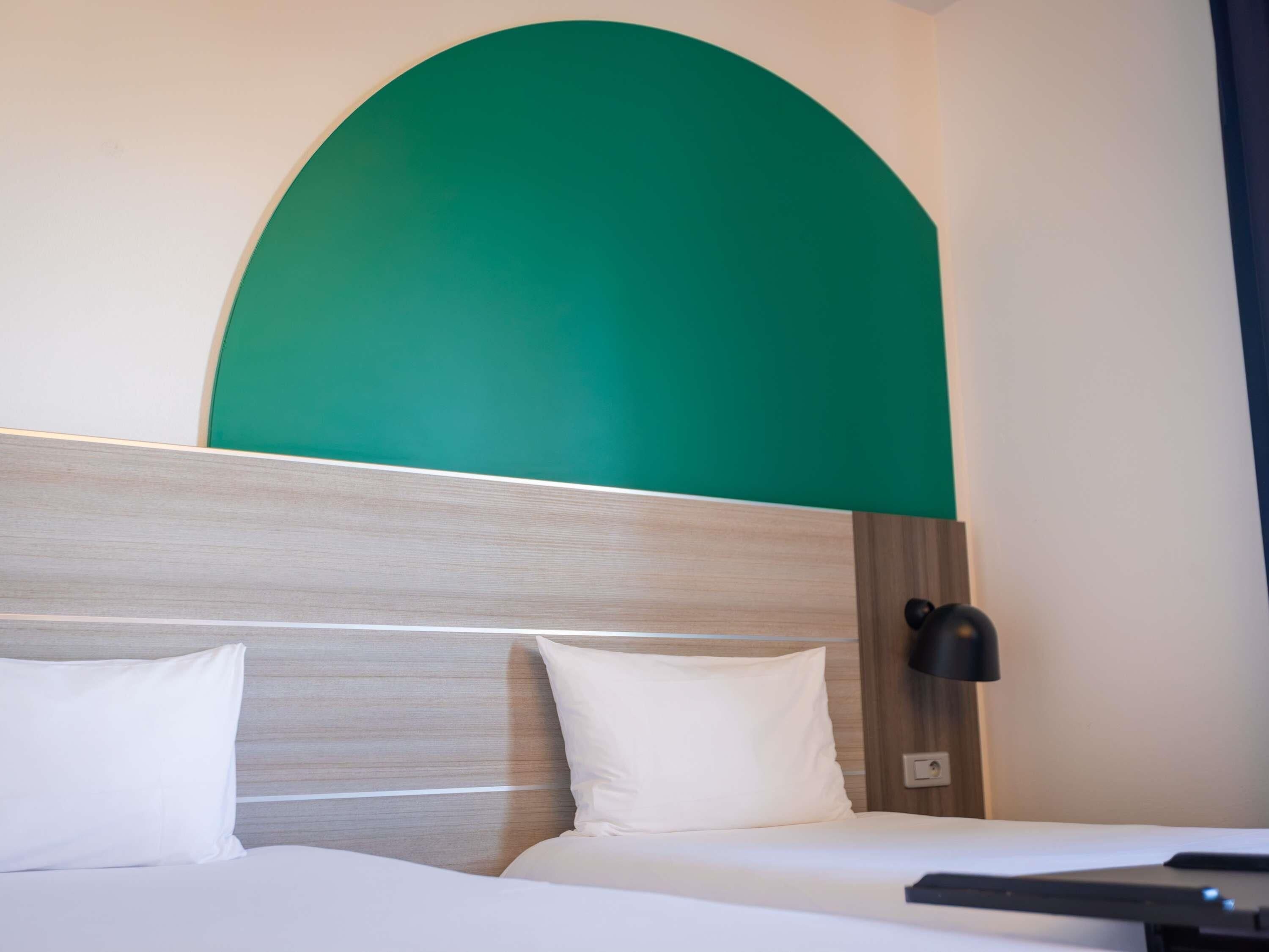 A hotel room with a bed, two pillows, a headboard with a wooden texture, and a green accent wall.