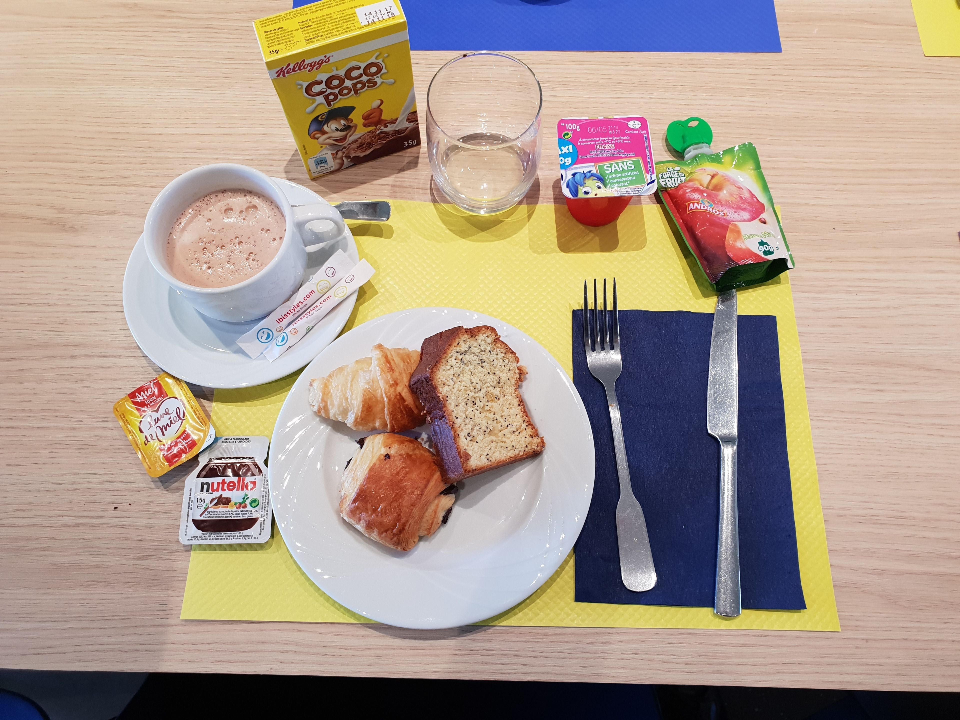 Un desayuno con una taza de café, croissants, un envase de Nutella, un vaso de agua y una caja de cereal Kellogg's Coco Pops.