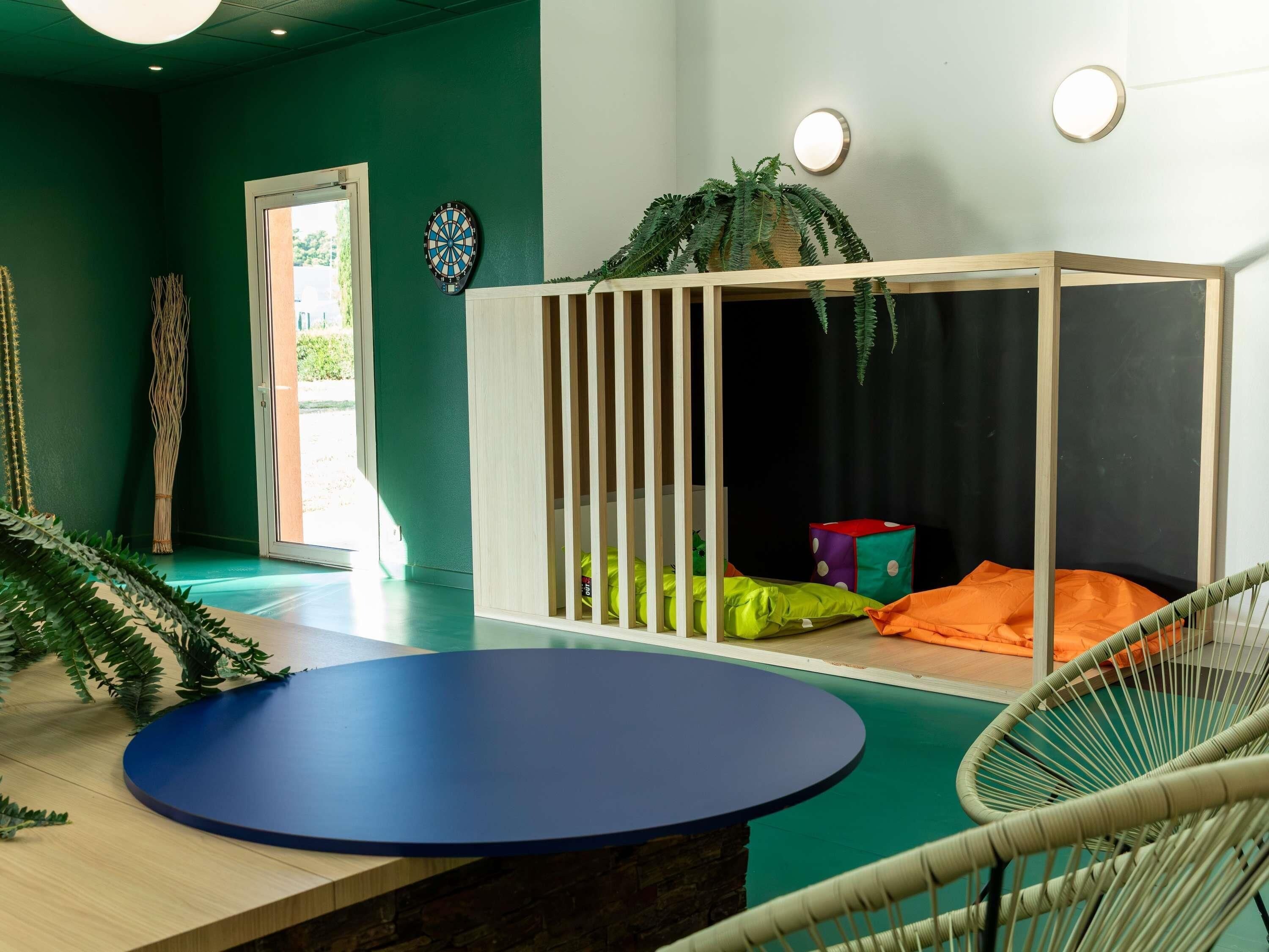 A room with a blue table, a white crib with colorful cushions, a dartboard, and a potted plant.