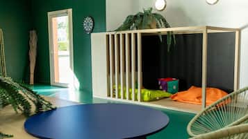 A room with a blue table, a white crib with colorful cushions, a dartboard, and a potted plant.