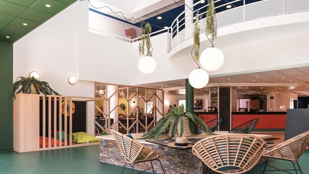 A modern indoor space with a central island table, wicker chairs, a green floor, and a mezzanine level with a railing.