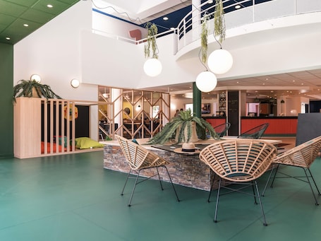 A modern indoor space with a central island table, wicker chairs, a green floor, and a mezzanine level with a railing.. ibis Styles Marseille Plan de Campagne