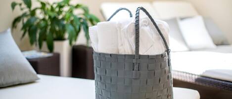 Una cesta gris trenzada con asas sobre una superficie blanca acolchada, probablemente en una habitación de hotel con una cama y una planta al fondo.
