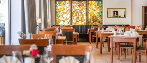 Un restaurante con varias mesas, sillas y un amplio ventanal de vidrio emplomado al fondo.
