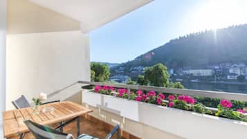A balcony with a wooden table and chairs, a flower vase, and a view of a river and hills.