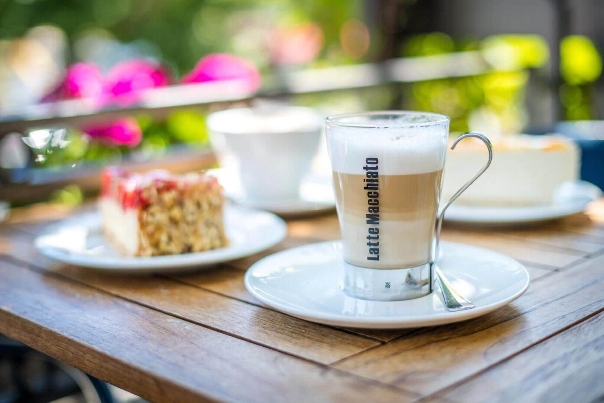 Una taza de latte macchiato sobre un platito blanco, una cuchara, una porción de pastel y una taza de café en una mesa de madera.
