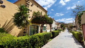 A pathway lined with neatly trimmed hedges and a palm tree, leading to a building with a beige facade and a red-tiled roof. - GF Isabel (Adeje)
