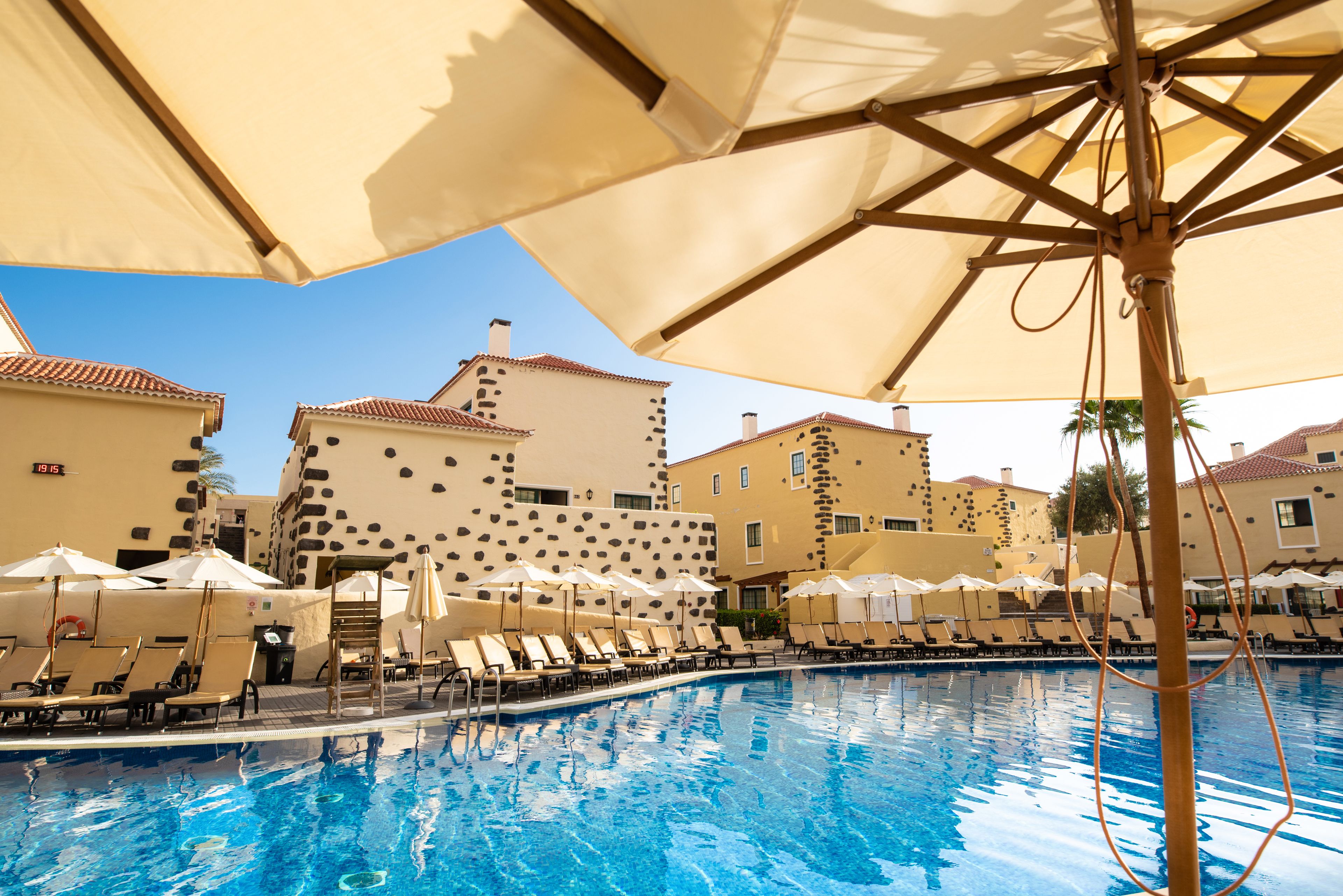Una piscina con sillas de descanso y sombrillas, rodeada de edificios con una fachada de piedra de diseño único.