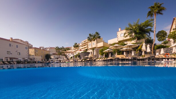 A clear blue pool with lounge chairs and umbrellas, surrounded by buildings and palm trees. - GF Isabel (Adeje)