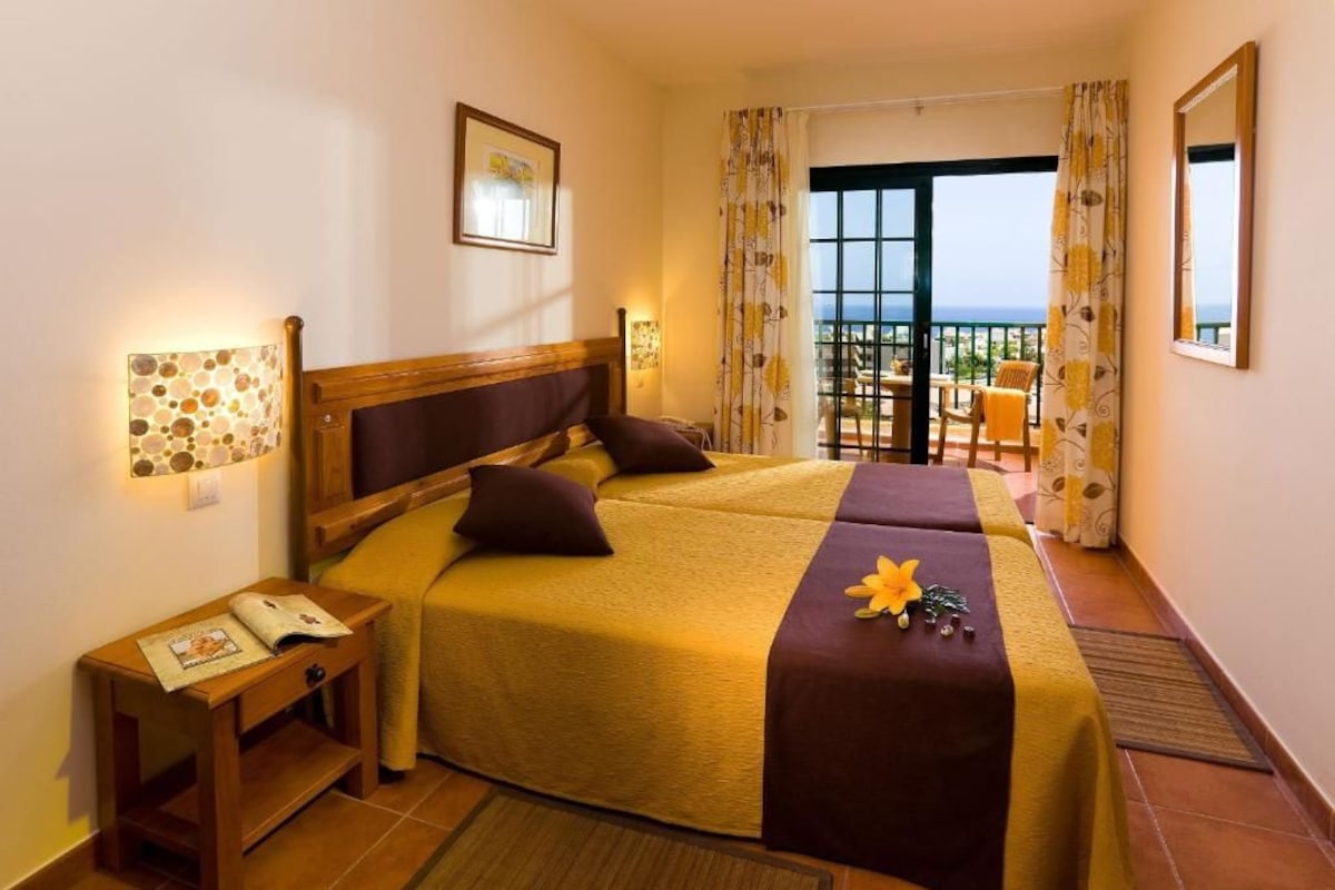 A hotel room with a large bed, a small bedside table, a nightstand, and a view of the balcony.