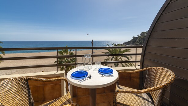 A balcony with wicker chairs, a table set with blue plates and silverware, and a clear view of the beach and ocean. - Almirall Apartaments (Lloret de Mar)