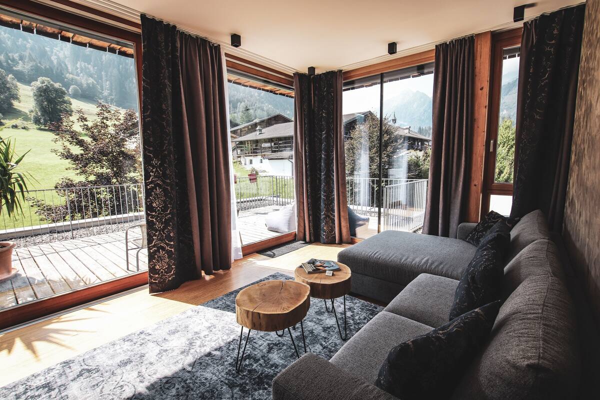 A modern living room with a large sofa, two round wooden side tables, and a view of a mountainous landscape through large windows.