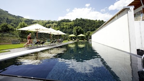 Una piscina con sillones de descanso y sombrillas, rodeada por un edificio blanco y un paisaje montañoso.