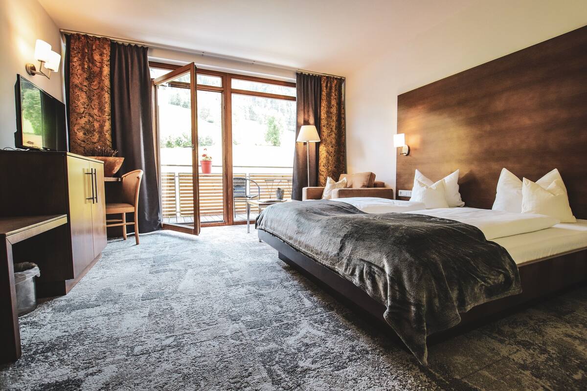 A hotel room with a large bed, a desk with a chair, a TV, and a balcony with a railing.