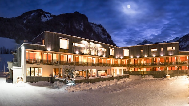Un hotel de varios pisos con el suelo cubierto de nieve, grandes ventanales y montañas al fondo.