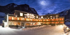 Un hotel de varios pisos con el suelo cubierto de nieve, grandes ventanales y montañas al fondo.