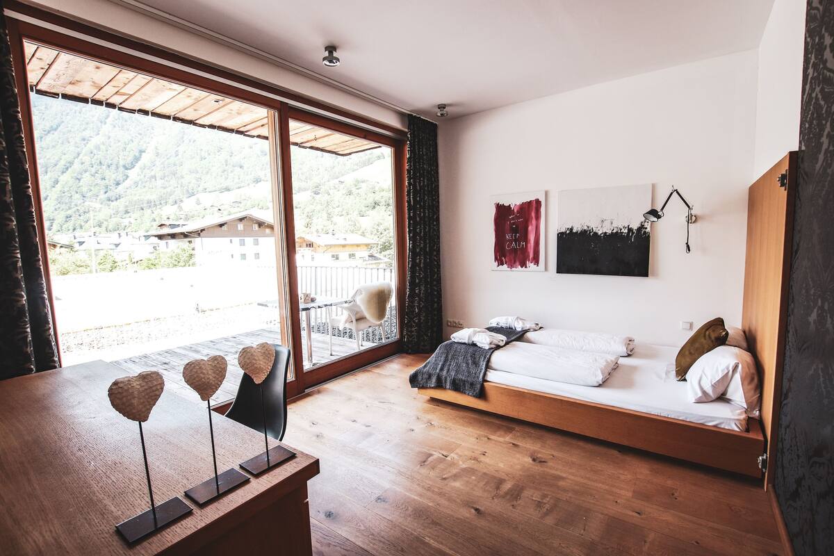 A bedroom with a wooden floor, a bed with pillows, a desk with heart-shaped decorations, and a view of a mountain through a large window.