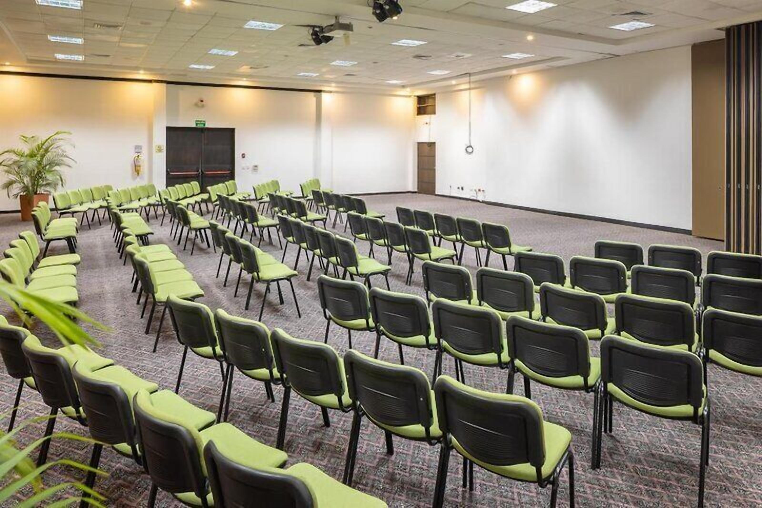 Uma sala de conferência com fileiras de cadeiras, uma tela de projetor e piso coberto de carpete.
