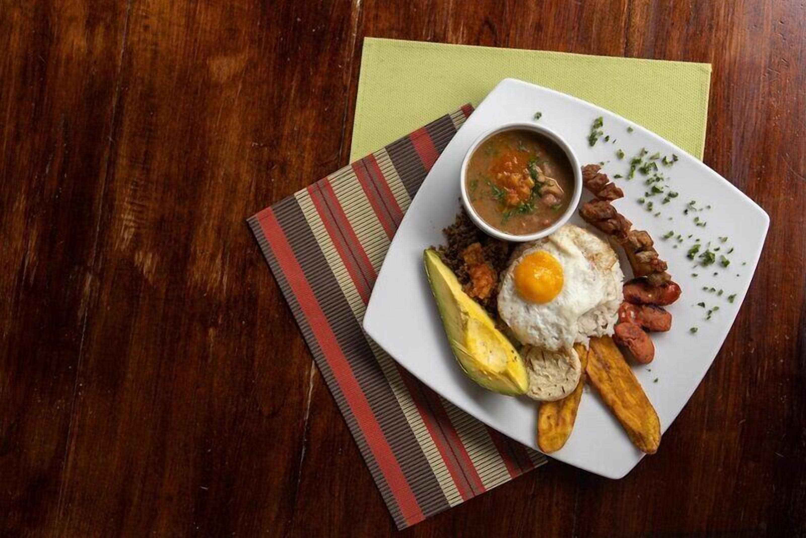 A plate of food with a fried egg, sausage, and a side of soup on a wooden table.